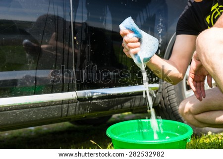 Close Up of Man Washing Car, Crouching Next to Black Car and Squeezing Soapy Blue Sponge Outdoors