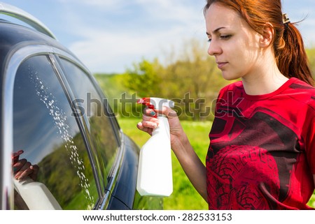 Woman Wearing Red Shirt Cleaning Car Windows with Spray Bottle Cleaner in Green Field on Bright Sunny Day