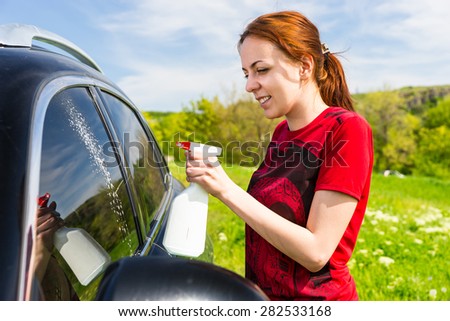 Woman Wearing Red Shirt Cleaning Car Windows with Spray Bottle Cleaner in Green Field on Bright Sunny Day