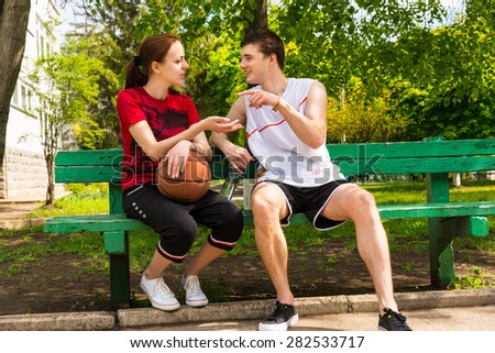 Young Man and Woman Wearing Athletic Clothing Sitting on Green Park Bench with Basketball Having a Conversation or Argument After the Game