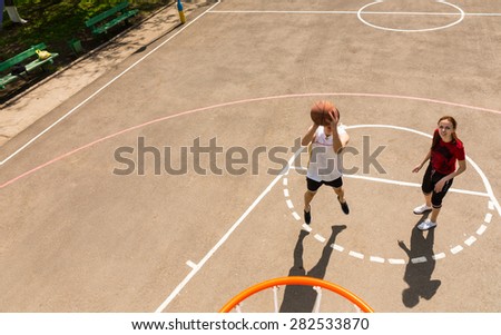 High Angle View from Backboard of Young Athletic Couple Playing Basketball - Woman Looks On While Man Takes Shot on Net on Outdoor Court on Sunny Day