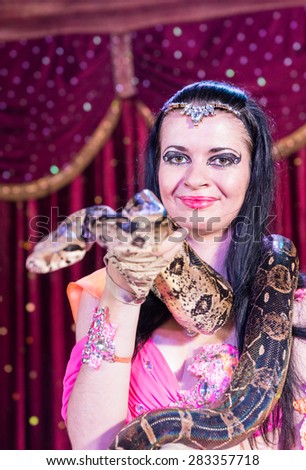 Head and Shoulders Close Up Portrait of Exotic Dark Haired Snake Charmer Wearing Large Snake Around Shoulders on Stage with Red Curtain