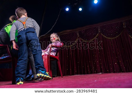 Low Angle View of Smiling Girl Wearing Clown Make Up Sitting in Red Chair on Stage with Boy and Man Looking On, Copy Space to Right of Image