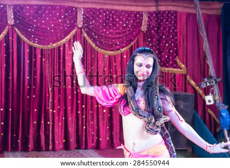 Waist Up of Exotic Dark Haired Belly Dancer with Large Snake Around Shoulders, Dancing on Stage with Red Curtain in Background