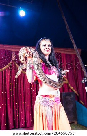 Exotic Dark Haired Belly Dancer Standing on Stage Holding Large Snake with Red Curtain in Background