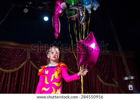 Cute girl with painted face and bright pink fantasy dress holding a bunch of gas colorful balloons during an artistic performance, in front of the dark red curtains