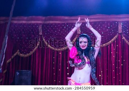 Waist Up of Exotic Dark Haired Belly Dancer with Large Snake Around Shoulders, Dancing with Arms Over Head on Stage with Red Curtain in Background