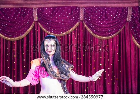 Waist Up of Exotic Dark Haired Belly Dancer with Large Snake Around Shoulders, Dancing on Stage with Red Curtain in Background