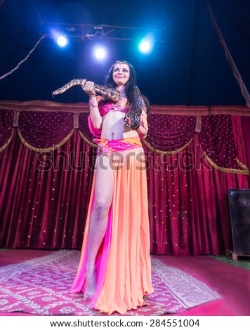 Full Length Low Angle View of Exotic Female Belly Dancer Wearing Bright Pink and Orange Costume with Large Snake Wrapped Around Shoulders Backlit by Bright Spotlights on Stage