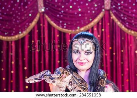 Head and Shoulders Close Up Portrait of Exotic Dark Haired Snake Charmer Wearing Large Snake Around Shoulders on Stage with Red Curtain