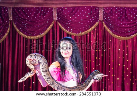 Waist Up of Exotic Dark Haired Belly Dancer Standing on Stage Holding Large Snake with Red Curtain in Background