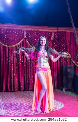 Full Length of Exotic Dark Haired Female Belly Dancer Wearing Bright Pink and Orange Costume with Large Snake Wrapped Around Shoulders on Stage with Red Curtain and Backlit by Bright Spotlights