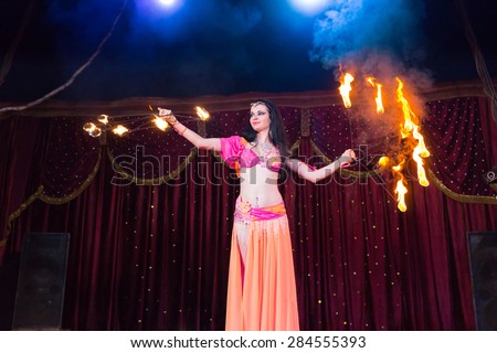 Exotic Dark Haired Fire Dancer Twirling Flaming Baton Apparatus on Stage Lit by Bright Spotlights