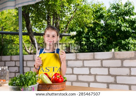 Young Girl Wearing Apron Holding Large Knife at Outdoor Table Covered with Fresh Vegetables
