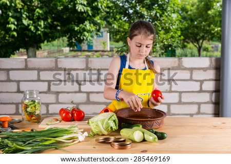 Young Girl Wearing Apron Chopping Fresh Vegetables for Canning or Preserving in Glass Jars, at Outdoor Table near Garden Brick Wall