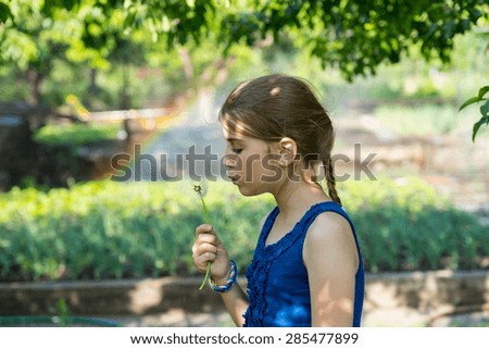 Pretty young girl blowing a dandelion clock in the garden with a rainbow on a sprinkler in the background making a colorful arc behind her