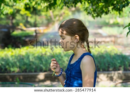 Cute genuine girl with long pony-tail and blue sleeveless top holding a fragile fuzzy dandelion while blowing on its seeds, outdoors in a green garden