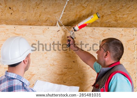 Construction Site Foreman Wearing White Hard Hat Holding Plans and Observing Worker Applying Caulking to Unfinished Wood Ceiling