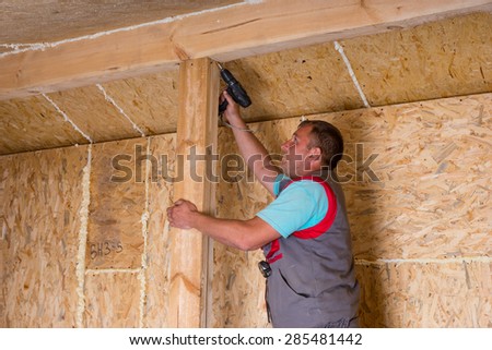 Male Construction Worker Builder with Cordless Drill Drilling Screw into Wooden Frame on Unfinished Home with Exposed Plywood Particle Board
