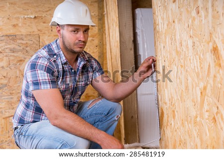 Male Construction Worker Builder Wearing White Hard Hat Measuring Door Frame with Tape Measure Inside Unfinished Home with Exposed Particle Plywood Boards