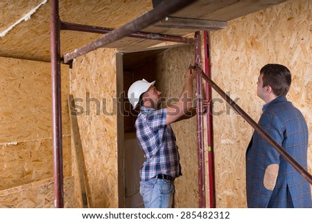 Architect in Suit Jacket Watching Construction Worker in Hard Hat Taking Measurements of Door Frame with Level Inside Unfinished New Home