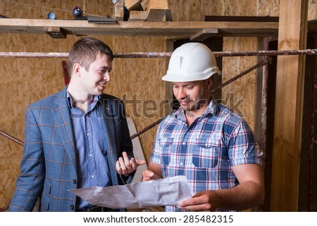 Young Male Architect and Construction Worker Foreman Inspecting Building Plans Together Inside Unfinished Building