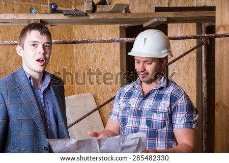 Young Male Architect and Construction Worker Foreman Inspecting Building Plans Together Inside Unfinished Building