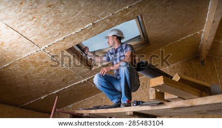 Low Angle View of Male Construction Worker Builder Crouching on Elevated Scaffolding near Ceiling and Inspecting Frame of Sky Light Window in Unfinished House with Exposed Particle Plywood Board