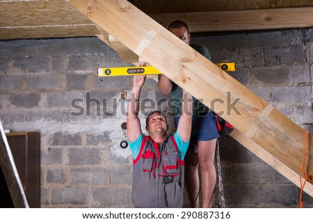 Two Men Working Together to Build Wooden Frame for Stairs Leading into Basement of Unfinished Home, Measuring Levelness of Steps Using Yellow Level Tool