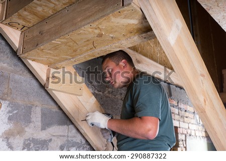 Male Construction Worker Builder with Cordless Drill Building Frame for Stairs in Basement of Unfinished Home with Exposed Cement Wall