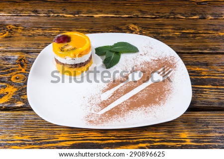 Close Up of Individual Decadent Fruit Cake Dessert Served on White Platter with Mint Leaf Garnish and Utensil Outlines Dusted in Cocoa on Wooden Table Surface