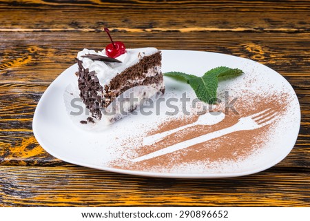 Slice of delicious chocolate cake garnished with a cherry and mint served on a platter with decorative detail of a fork and spoon in chocolate powder