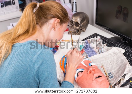 Close Up of Young Blond Artistic Woman Painting Colorful Details Onto Clay Tribal Mask While Sitting at Computer Desk