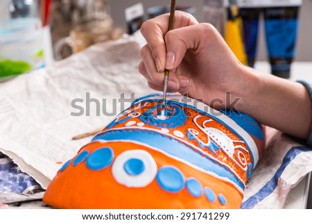 Artist painting the colorful decorative headdress on a handmade mask in a concept of creativity and handicraft work, close up view of her hand and paintbrush