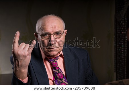 Elderly man making a horns gesture depicting heavy metal rock music or the sign of the devil, seated head and shoulders portrait on a dark background with copyspace