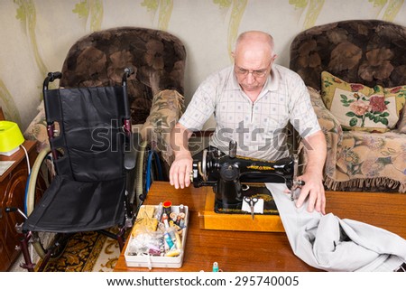 High Angle View of Senior Man at Home Using Old Fashioned Manual Sewing Machine to Mend Pants, in Living Room with Wheelchair Close By