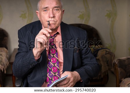Close up Middle Aged Bald Businessman Holding Pen to Corner of his Mouth and Small Notes, Looking at the Camera Seriously
