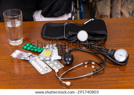 High Angle View of Various At-Home Blood Pressure Medical Supplies on Table - Glass of Water, Various Pills in Foil Blister Packs, Stethoscope and Blood Pressure Cuff with Gauge