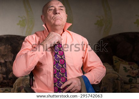 Handsome senior man is trying on a beautiful tie sitting in his living room with very serious face wearing a stylish shirt and several ties on his hand