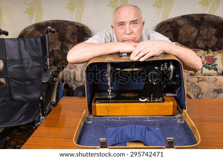 Lonely Old Bald Man Leaning on his Sewing Machine in a Carrying Case On Top of the Table and Looking at the Camera.