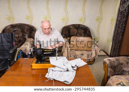 High Angle View of Senior Man Using Old Fashioned Manual Sewing Machine to Mend Pants at Home in Living Room with Wheelchair Close By