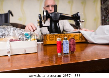 Senior Man Using Old Fashioned Manual Sewing Machine to Mend Clothing with Focus on Colorful Spools of Thread in Foreground