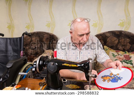 Senior Man Working On Needle Point Wall Hanging and Seated in front of Old Fashioned Manual Sewing Machine in Living Room at Home