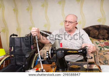 Senior Man with Measuring Tape Sitting in front of Old Fashioned Manual Sewing Machine Sitting Beside Wheelchair at Home in Living Room