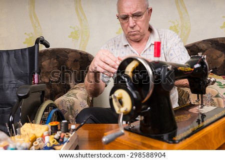 Senior Man Wearing Eyeglasses and Looking Down at Hands While Sitting in Living Room at Old Fashioned Sewing Machine