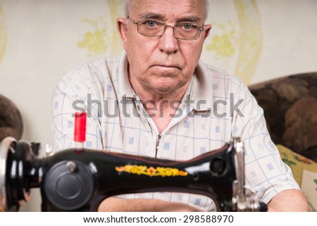 Close Up of Serious Senior Man Wearing Eyeglasses Using Old Fashioned Sewing Machine at Home in Living Room