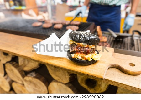 Gourmet Burger Served Alone on Wooden Cutting Board with Napkins on Counter of Food Stall with Person Cooking Patties on Grill in Background