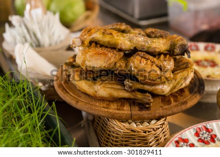 Roasted Grilled Chicken Legs Stacked on Wooden Platter and Served on Table Surrounded by Other Dishes
