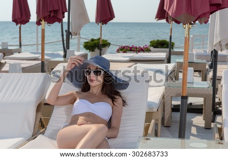 Fashionable young woman sunbathing in a bikini as she relaxes at a coastal resort on a recliner chair wearing trendy sunglasses and sunhat