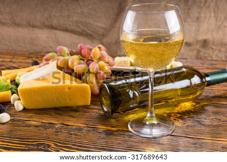 Gourmet Food Still Life - Glass of White Wine on Rustic Wooden Table with Fallen Bottle and Variety of Cheeses and Grapes